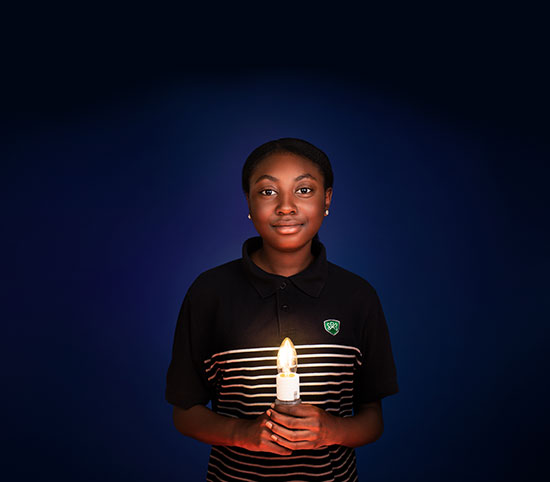 Against a dark blue background, a dark-skinned girl in a school uniform holds a light bulb that illuminates her face.
