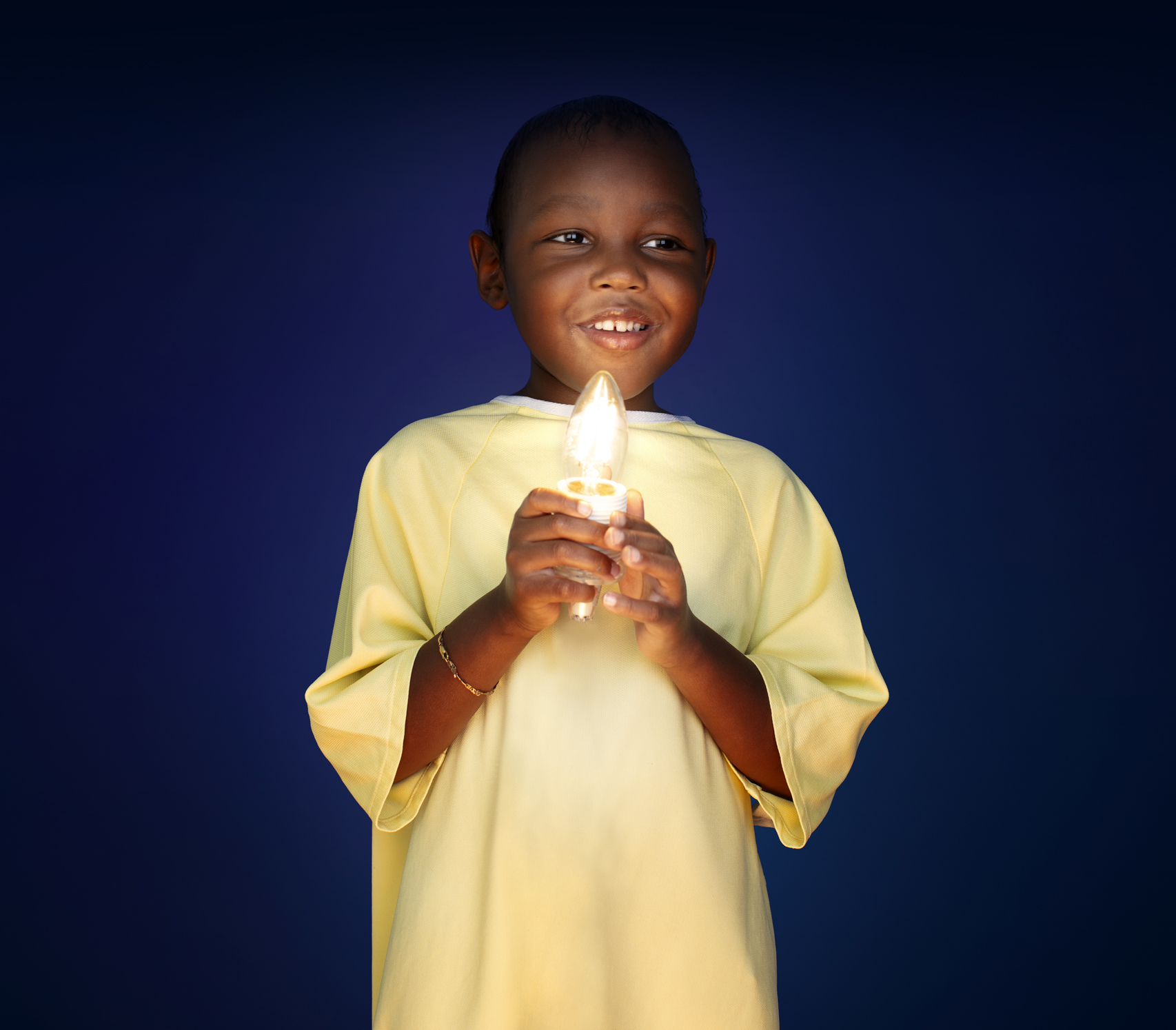 Five-year-old Eli-Noah, dressed in a yellow hospital gown, holds up a glowing candle-shaped light in his hands.
