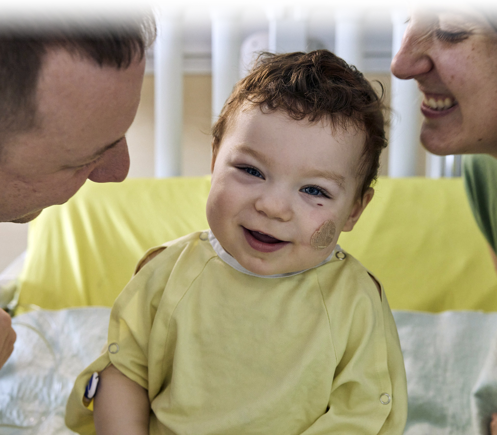 Julien, 16 mois, vêtu d'une jaquette jaune, est assis sur son lit d'hôpital et sourit tendrement aux cotés de ses parents.