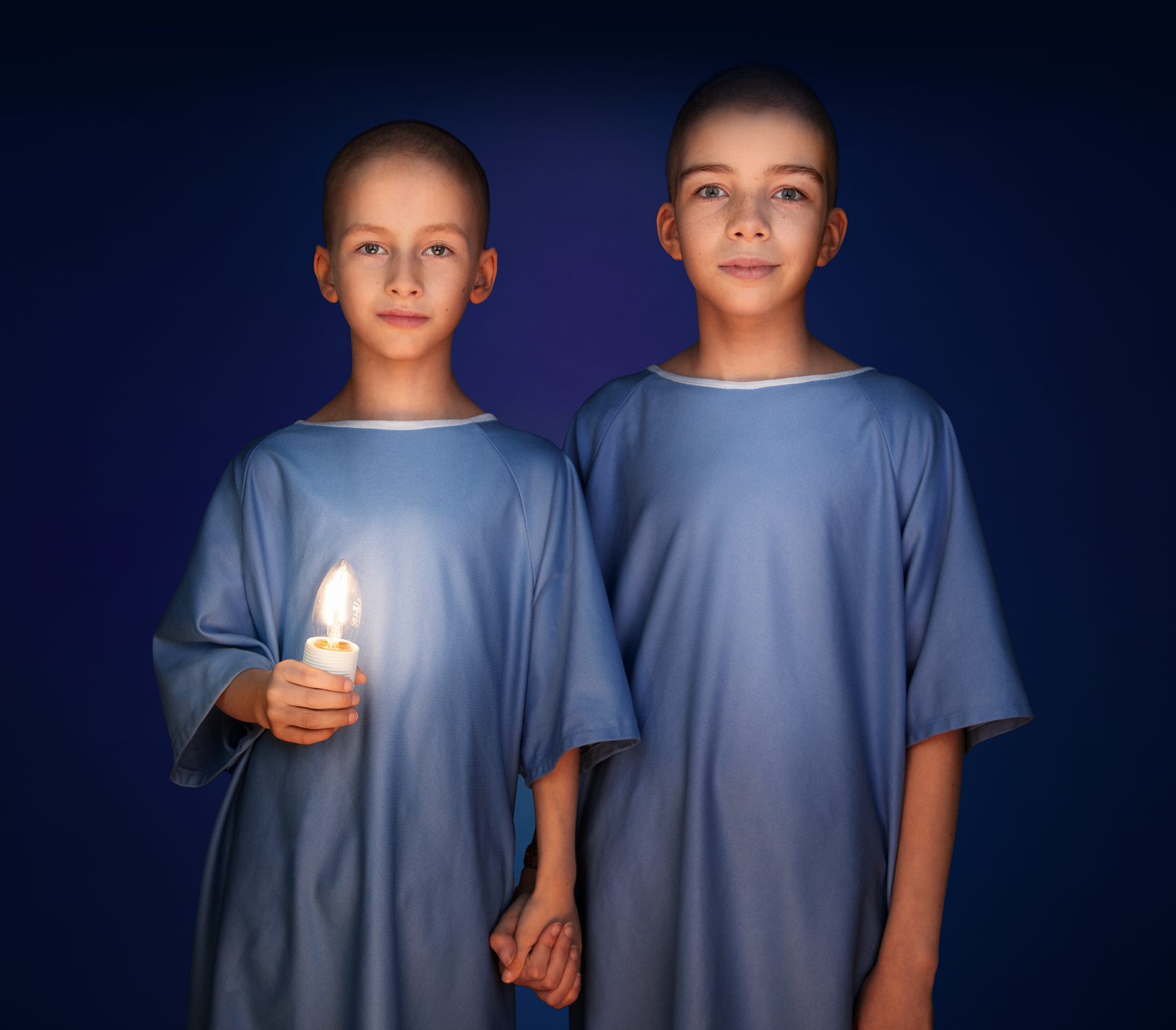 Siblings Jules and Lou, both wearing blue hospital gowns, stand side by side, smiling and holding hands. Jules has a glowing candle-shaped light in his other hand.