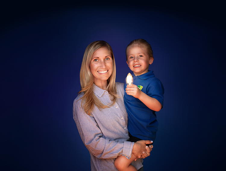 Against a dark blue background, a mother carries her little boy in her arms who is holding a light bulb that illuminates his face.