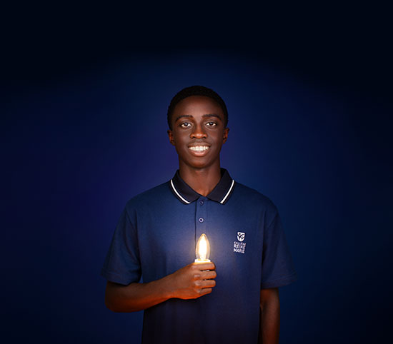 Sur fond bleu foncé, un adolescent en uniforme scolaire à la peau foncée tient une ampoule qui illumine son visage souriant.