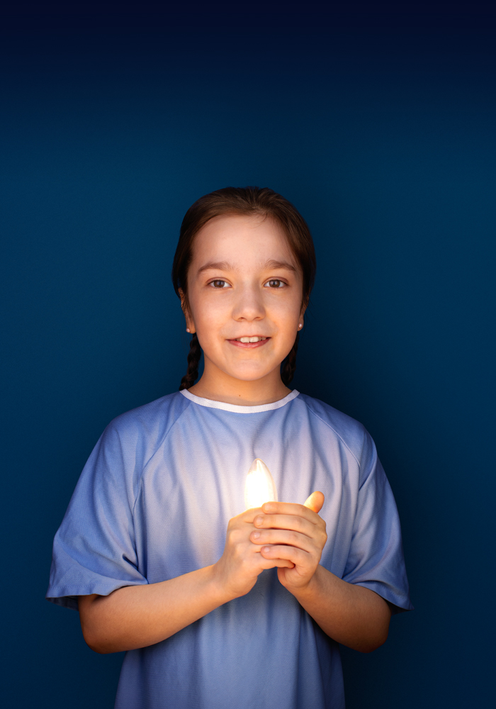 Gabrielle, 12 ans, porte une jaquette d’hôpital bleue, sourit et tient une ampoule allumée dans ses mains.