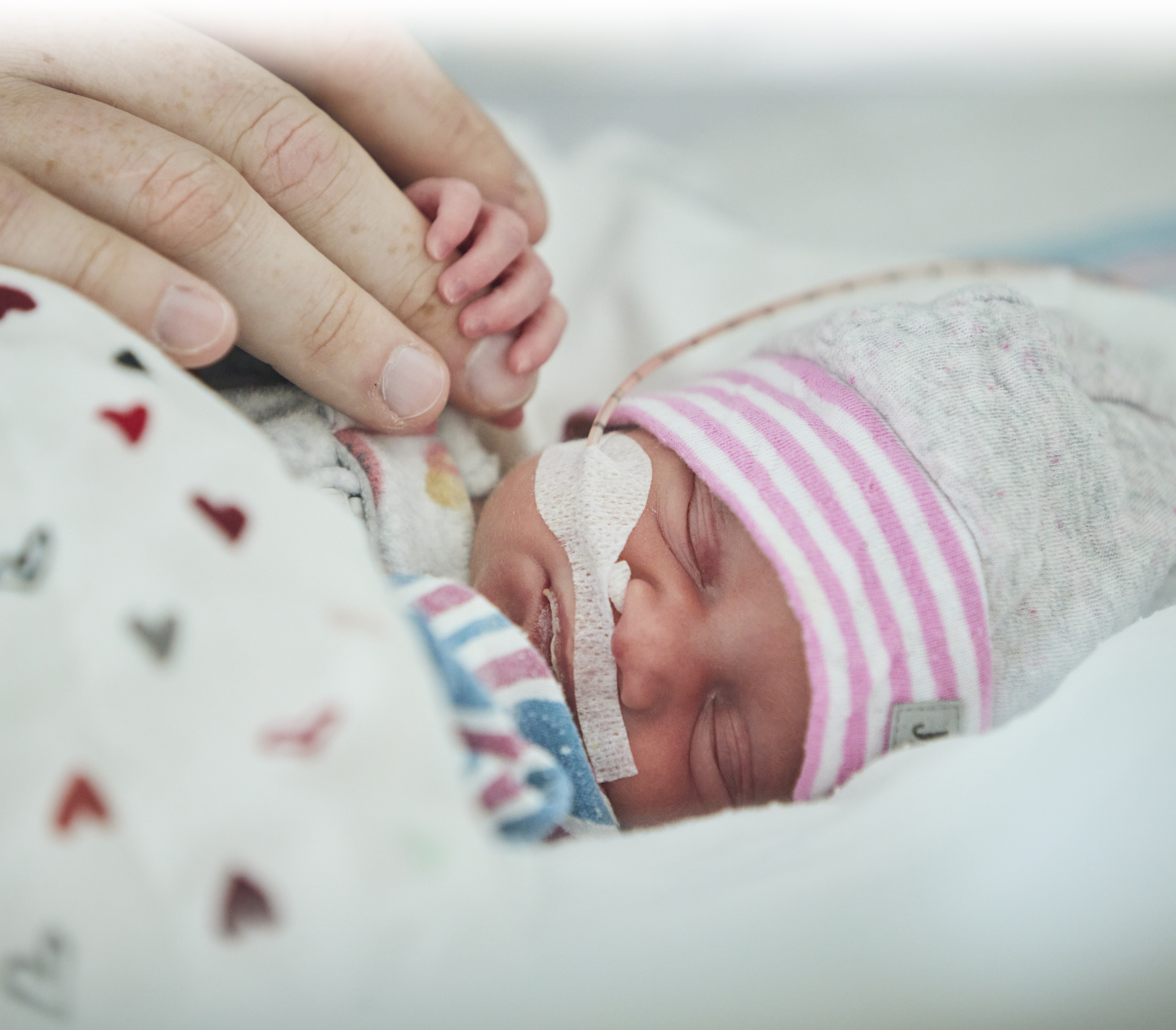 Elyrose, un bébé à l'unité de néonatalogie du CHU Sainte-Justine, est couchée, les yeux fermés, et tient le doigt de son papa avec minuscule main.