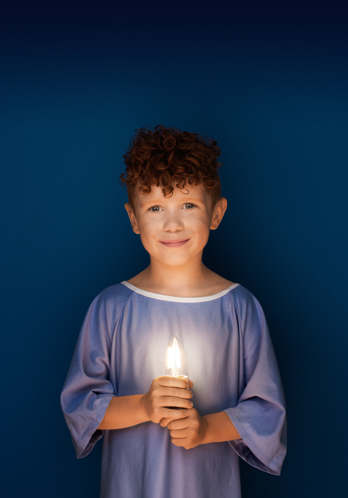 Adam, 6 ans, porte une jaquette d’hôpital bleue, sourit et tient une ampoule allumée dans ses mains.