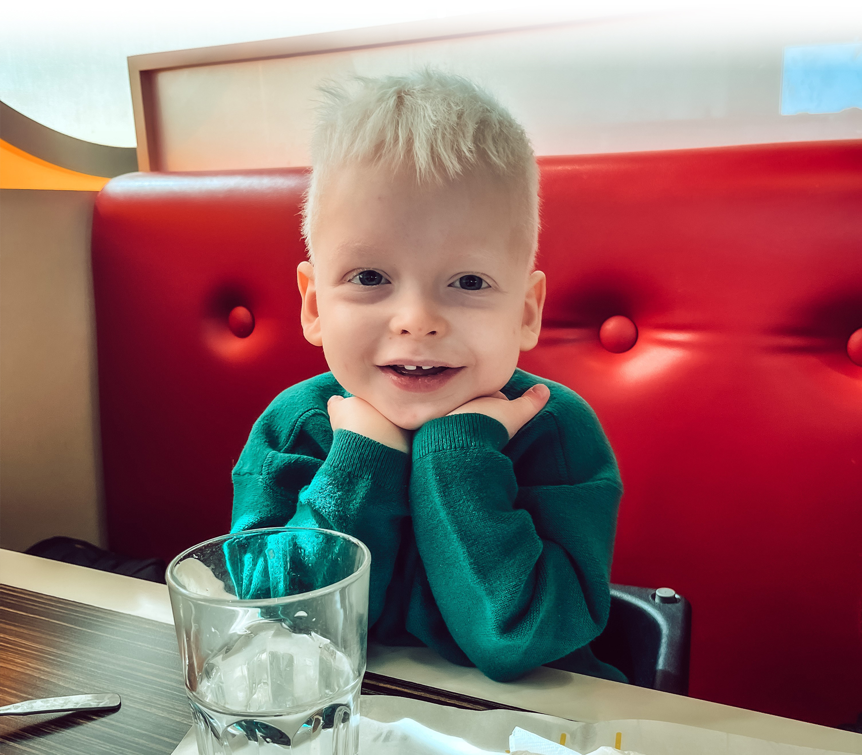 A beaming four-year-old Milan, sitting on a red upholstered restaurant bench with his elbows propped up on the table in front of him.