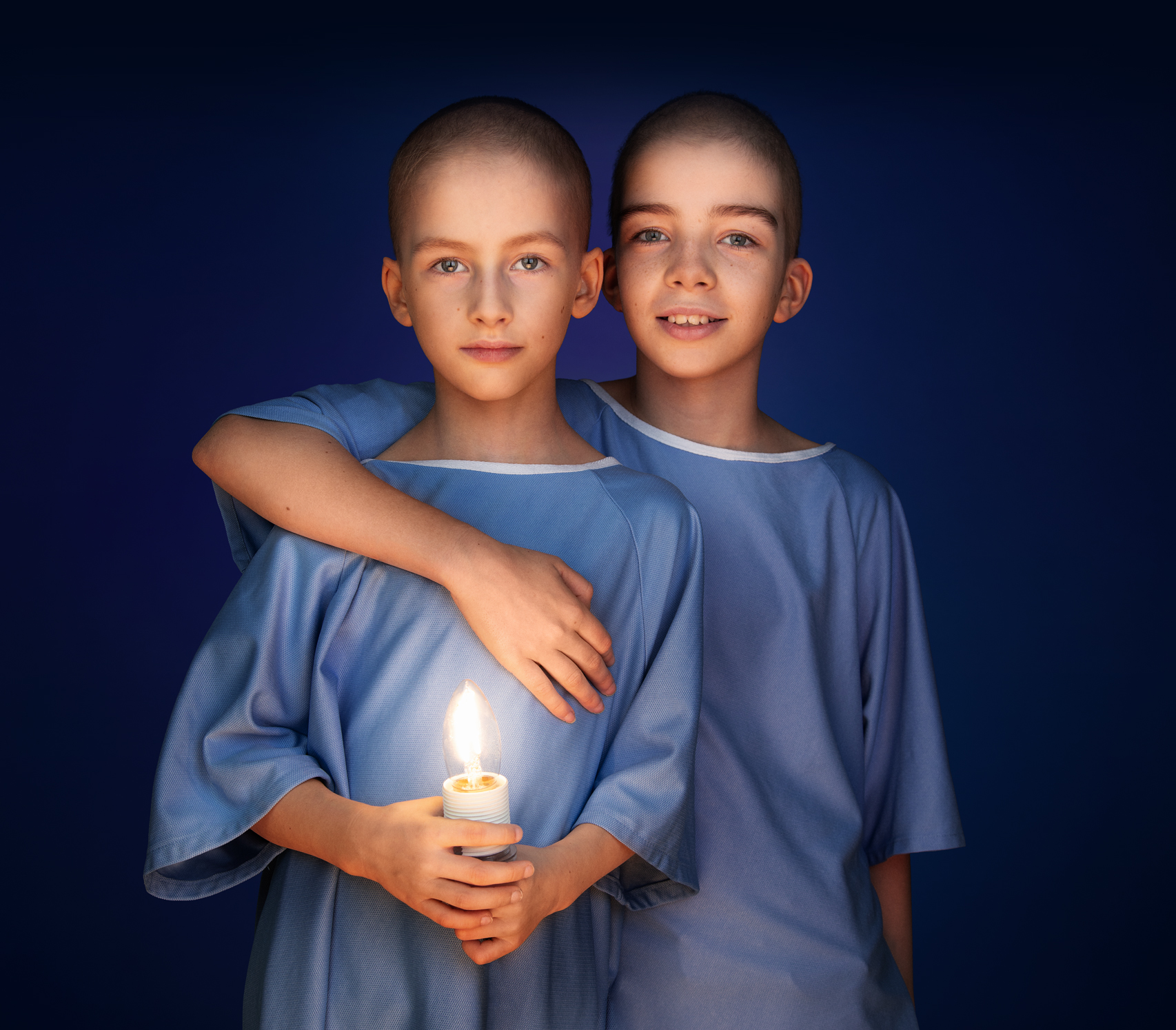 Sister Lou stands behind brother Jules with her arm wrapped around him. Both are wearing blue hospital gowns. Jules is holding a glowing candle-shaped light in his hands.